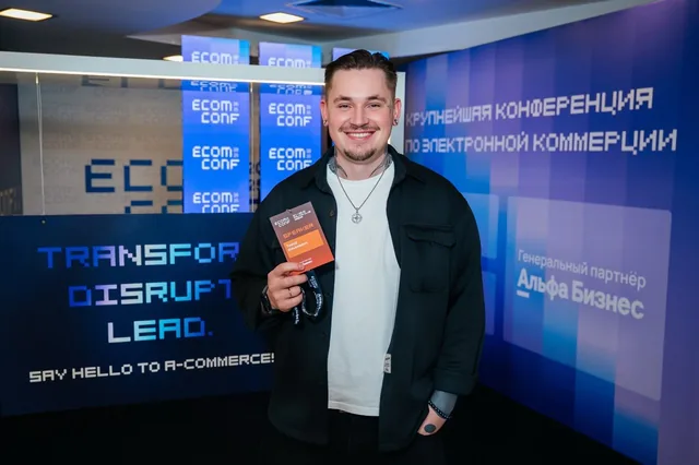 Тимур улыбается в холле Ecom Conf и держит бейдж спикера на фоне стенда конференции.